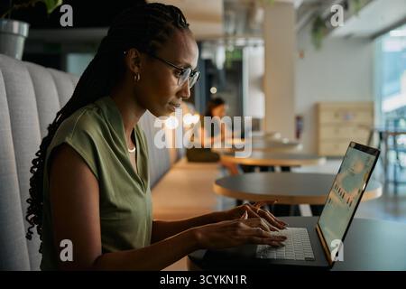 Vue latérale de la jeune femme d'affaires noire concentrée tapant sur l'ordinateur portable assis au bureau dans le salon de bureau Banque D'Images