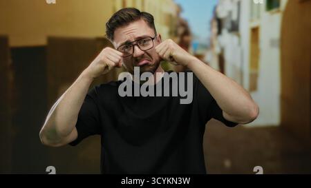 Jeune homme avec la barbe et les lunettes prétendant pleurer dans un cadre extérieur de rue de ville mettant en valeur l'expression ludique et l'émotion comique. Banque D'Images
