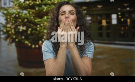 Jeune femme hispanique en chemise bleue souffle doucement baiser avec les deux mains sur la rue ensoleillée ; affection amour. Banque D'Images