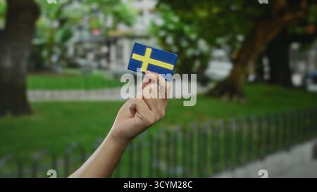 Main tenant le drapeau suédois à l'extérieur dans un cadre de parc, avec un homme caucasien visible, représentant la culture et l'identité suédoises. Banque D'Images