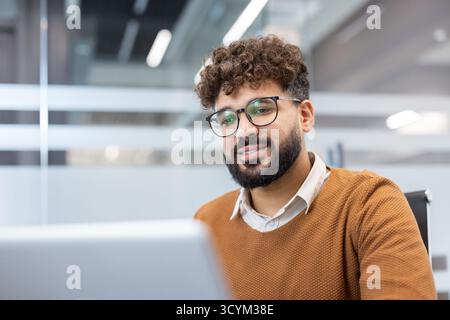 Jeune homme professionnel portant des lunettes et une barbe souriant avec confiance, travaillant sur un ordinateur portable dans un bureau moderne, incarnant le dévouement et le succès de l'entreprise Banque D'Images