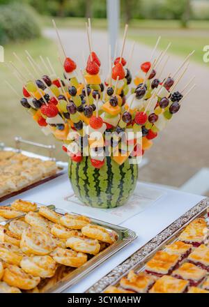 Une vif exposition de brochettes de fruits disposées dans un vase de pastèque, entouré de plateaux de pâtisserie. La combinaison de fraises, de raisins et de melon Banque D'Images