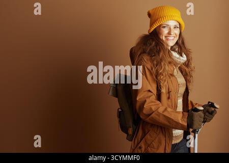 Une femme souriante dans les années 30, portant un bonnet jaune, une veste à capuche marron et un pull à motifs, pose avec des bâtons de trekking et un sac à dos vert, prêt Banque D'Images
