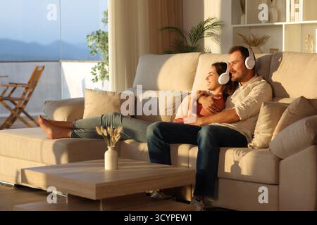 Couple heureux écoutant de la musique avec des écouteurs regardant loin assis sur un canapé à la maison Banque D'Images