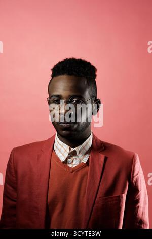 Portrait de jeune homme africain à la mode portant des lunettes regardant la caméra tout en se tenant dans un faible éclairage sur fond rouge Banque D'Images