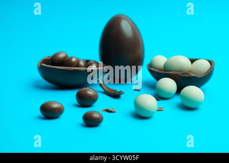 Oeuf entier à côté de deux moitiés craquelées révélant des bonbons au chocolat noir et blanc sur un fond bleu vif - idéal pour la photographie culinaire des fêtes. Banque D'Images