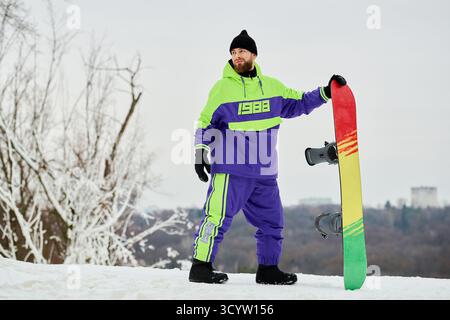 Un homme barbu se prépare au snowboard sur une pente hivernale tout en portant des vêtements colorés. Banque D'Images