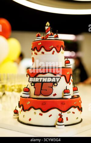 Gâteau festif à trois couches décoré avec fondant rouge et blanc, une grande bougie, et le chiffre 15. Cadre de célébration d'anniversaire avec ballons. Banque D'Images