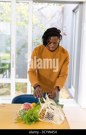 Homme afro-américain adulte moyen dans le pull moutarde déballage des produits du sac en maille sur la table de cuisine Banque D'Images