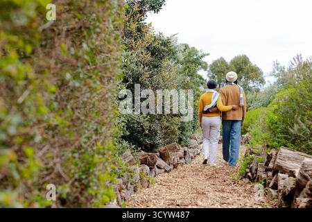 Couple afro-américain marchant bras dans bras entre des haies taillées et des bûches empilées sur le chemin des copeaux de bois Banque D'Images