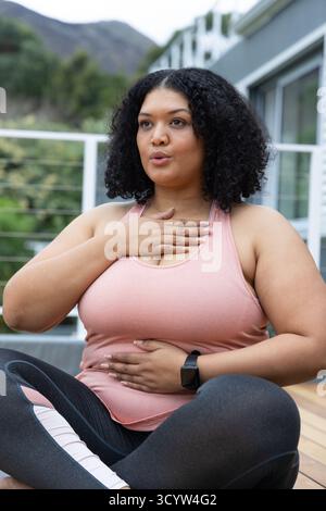 Femme afro-américaine assise sur le pont en bois pratiquant l'exercice de respiration dans le haut rose, smartwatch Banque D'Images