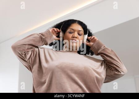 Femme afro-américaine en sweat-shirt beige plaçant des écouteurs sans fil tout en se tenant debout à la maison Banque D'Images