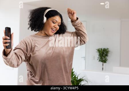 Femme afro-américaine dansant en sweat-shirt et écouteurs à la maison tenant smartphone, espace copie Banque D'Images