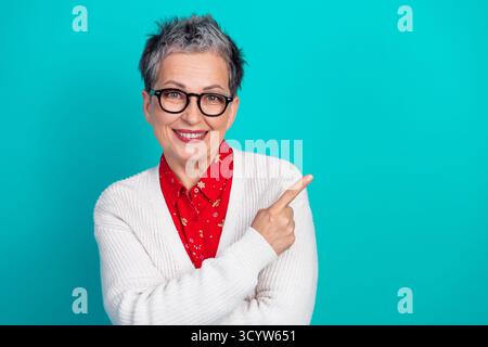 Femme mature souriante avec des lunettes dans une chemise rouge et un cardigan blanc pointe vers la droite sur un fond bleu sarcelle lumineux exprimant l'humeur des vacances Banque D'Images