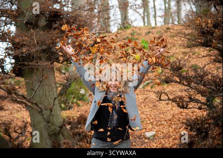 Une femme blonde de 40 ans vêtue d'élégants vêtements gris jette des feuilles d'automne vers le haut, pleines de joie de vivre. Une forêt automnale à feuilles caduques peut être vue floue Banque D'Images