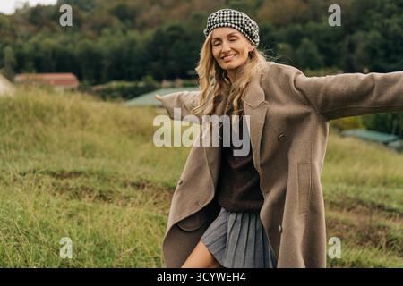 Heureuse femme souriante dans un béret et un manteau se promène à travers la campagne en automne. Banque D'Images