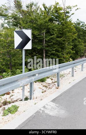 Un panneau routier audacieux pointant vers la droite se dresse près d'un garde-corps métallique, avec des arbres verts denses et du gravier rocheux en toile de fond. La scène transmet l'avertissement, gu Banque D'Images