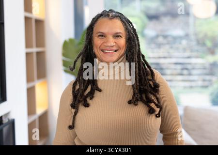 Femme afro-américaine debout dans le salon souriant mettant en évidence les casiers d'étagères et la plante d'intérieur Banque D'Images
