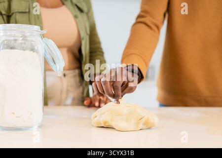 Couple afro-américain pétrissant la pâte sur le comptoir blanc dans la cuisine avec pot de farine et montre-bracelet Banque D'Images