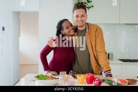 Couple diversifié souriant et préparant la salade sur planche à découper avec des poivrons, concombre dans la cuisine Banque D'Images