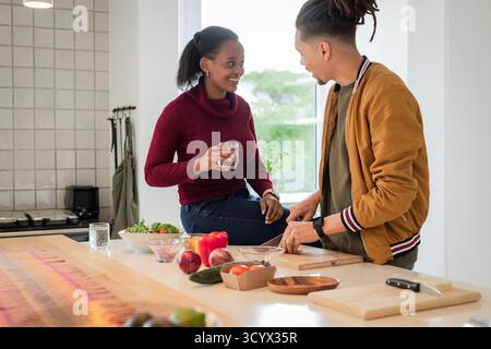 Couple diversifié préparant la salade sur l'îlot de cuisine avec le verre de planche à découper en bois de l'eau et des bols Banque D'Images
