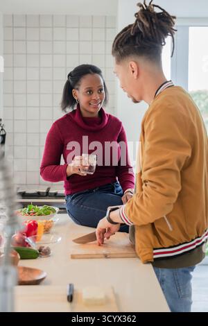 Couple diversifié hachant des légumes sur une planche à découper et sirotant de l'eau près du bol à salade dans la cuisine Banque D'Images