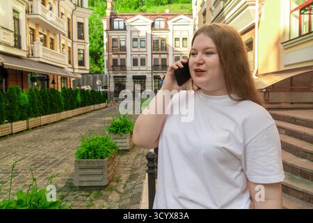 Femme de grande taille confiante utilisant son téléphone portable pour une conversation tout en se tenant debout dans une rue pavée entourée de bâtiments européens classiques Banque D'Images
