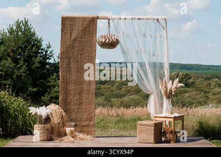 Une belle arche de cérémonie de mariage en plein air avec une toile de fond de jute, des rideaux blancs transparents, des accents d'herbe de pampa et des meubles en bois dans un s. Banque D'Images