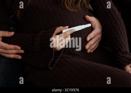 Un couple aimant tient doucement un test de grossesse positif sur la bosse de bébé d'une femme, partageant un moment tendre de joie et d'anticipation. tricots marron et s Banque D'Images