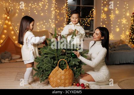 Mère et enfants décorent une petite épicéa dans une maison confortable avec des lumières scintillantes, idéal pour les campagnes de vacances, les cartes et les annonces saisonnières. Banque D'Images