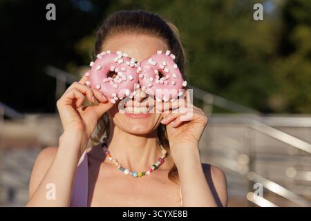 Joyeuse femme couvre les yeux avec des beignets givrés roses surmontés de guimauves portrait de dessert ludique à l'extérieur dans la lumière chaude du soleil Banque D'Images