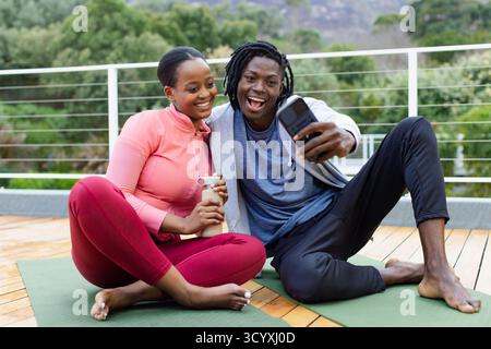 Couple afro-américain assis sur des tapis de yoga sur une terrasse en bois tenant une bouteille d'eau et un smartphone Banque D'Images