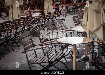 Rangées de chaises en bois vides et tables rondes dans un café extérieur fermé à Ljubljana, Slovénie, avec des parasols pliés et un trottoir humide suggérant off-se Banque D'Images