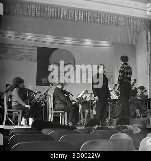 Cette photo d'archive de 1966 de Sloviansk, RSS d'Ukraine, montre un orchestre de chambre en train de répéter ou de se produire sur scène. Le cadre est probablement un Palais de la culture. La toile de fond est dominée par un grand portrait de Vladimir Lénine et des slogans soviétiques ('nous disons Lénine, nous entendons le Parti'). Des musiciens, principalement des cordes, jouent sous la direction d'un chef d'orchestre sur un podium. Certaines images montrent un chanteur masculin en train de jouer. Cette scène capture parfaitement la vie culturelle et artistique dans une ville soviétique, un document du Donbass paisible Banque D'Images