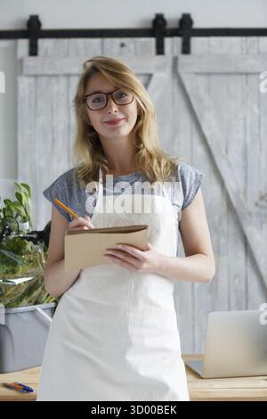 Jeune femme fleuriste avec des lunettes tenant le cahier en magasin Banque D'Images