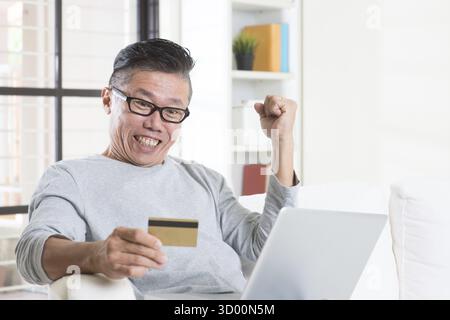 Portrait de l'homme asiatique mature des années 50 gagnant des prix tout en utilisant l'Internet informatique faisant le paiement en ligne avec carte de crédit, assis sur le canapé à la maison Banque D'Images