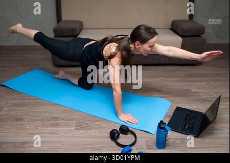 Femme pratiquant pilates pour un dos sain sur tapis de yoga à la maison, suite à l'entraînement en ligne sur ordinateur portable, avec haltères, bande de résistance et rouleau de mousse nea Banque D'Images
