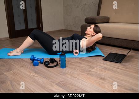Femme sur tapis de yoga à la maison effectuant des exercices avec rouleau en mousse pour un dos sain, suite à la formation de fitness en ligne sur ordinateur portable, bien-être et LIF actif Banque D'Images