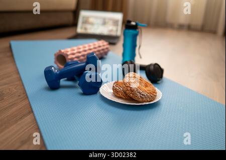 Appareil de fitness avec haltères, rouleau en mousse, bouteille d'eau, écouteurs et plaque de biscuits sur tapis de yoga à la maison, ordinateur portable en arrière-plan, concept de onl Banque D'Images