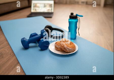 Appareil de fitness avec haltères, rouleau en mousse, bouteille d'eau, écouteurs et plaque de biscuits sur tapis de yoga à la maison, ordinateur portable en arrière-plan, concept de onl Banque D'Images
