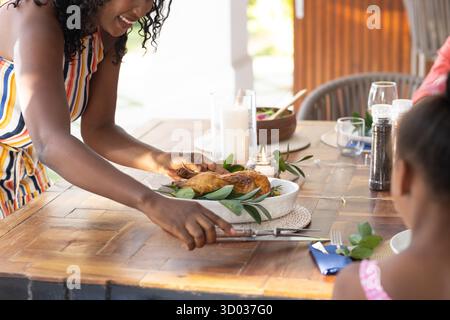 Servant du poulet rôti au dîner familial, femme afro-américaine souriant chaleureusement. célébration, homecooked, rassemblement, repas, bonheur, tradition || autorisation du modèle Banque D'Images