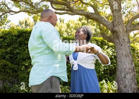 Couple senior dansant joyeusement ensemble dans le jardin ensoleillé sous l'arbre. Personnes âgées, bonheur, plein air, retraite || modèle libéré Banque D'Images