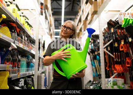 Une femme est engagée dans le shopping, choisissant joyeusement un arrosoir vert vif au milieu d'une gamme colorée d'outils de jardinage et d'articles ménagers. || autorisation du modèle Banque D'Images