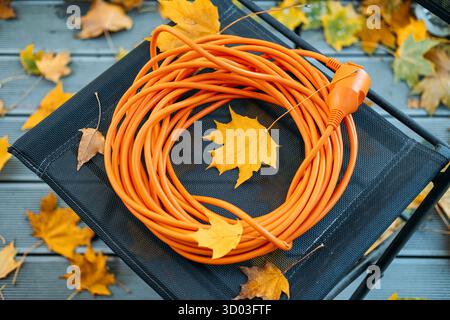 Rallonge orange vif enroulée sur un tabouret en maille noire parmi les feuilles d'automne dispersées sur un pont en bois. Sécurité technique extérieure, préparation saisonnière Banque D'Images