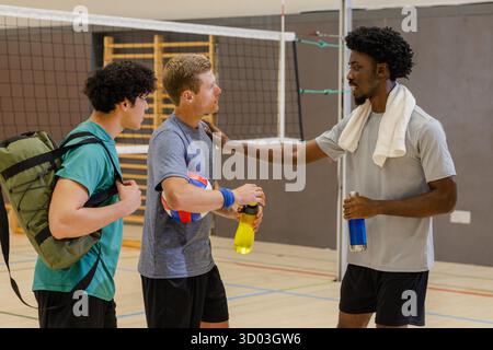 Divers coéquipiers masculins se reposant sur le terrain de volley-ball de gymnastique par filet tenant le volley-ball et des bouteilles d'eau. Athlétique, travail d'équipe, fitness, vêtements de sport, mouvement, camaraderie, énergique || modèle libéré Banque D'Images