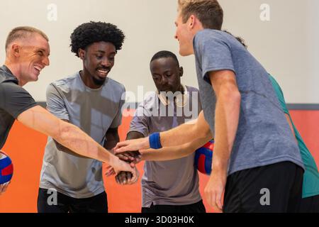 Divers coéquipiers masculins empilant les mains dans une salle de sport tenant des balles de volley et portant des écouteurs. Athlètes, travail d'équipe, entraînement, dynamique, camaraderie, fitness, compétition || autorisation du modèle Banque D'Images