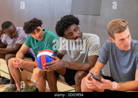 Divers coéquipiers masculins de volley-ball se reposant sur un banc en bois dans la salle de gym tenant le volley-ball et le smartphone. Athlètes, travail d'équipe, décontracté, sportif, fitness, camaraderie, compétition || libération du modèle Banque D'Images