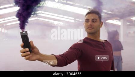 Homme portant une chemise rouge libérant de la fumée violette épaisse avec un appareil dans l'entrepôt, icônes sociales flottantes Banque D'Images
