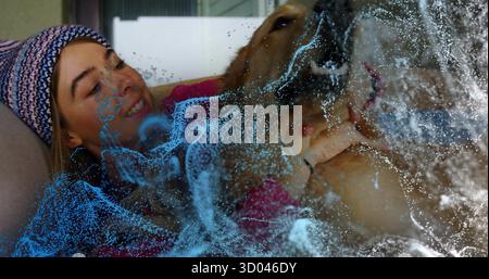 Femme embrassant dans un bonnet et un pull rouge tenant Golden retriever sur le canapé, avec des particules numériques Banque D'Images