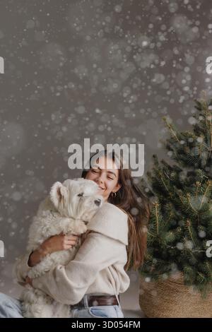 Jeune femme embrasse Westie moelleux près de Evergreen comme la neige tombe dans le studio, confortable portrait de style de vie de vacances d'hiver Banque D'Images